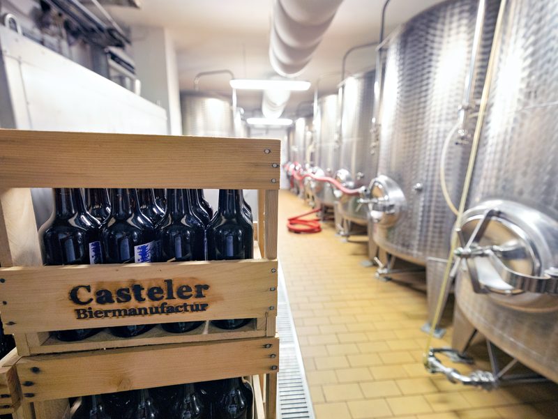 Holzkiste mit dem Schriftzug Casteler Biermanufactur vor einer Reihe Edelstahltanks